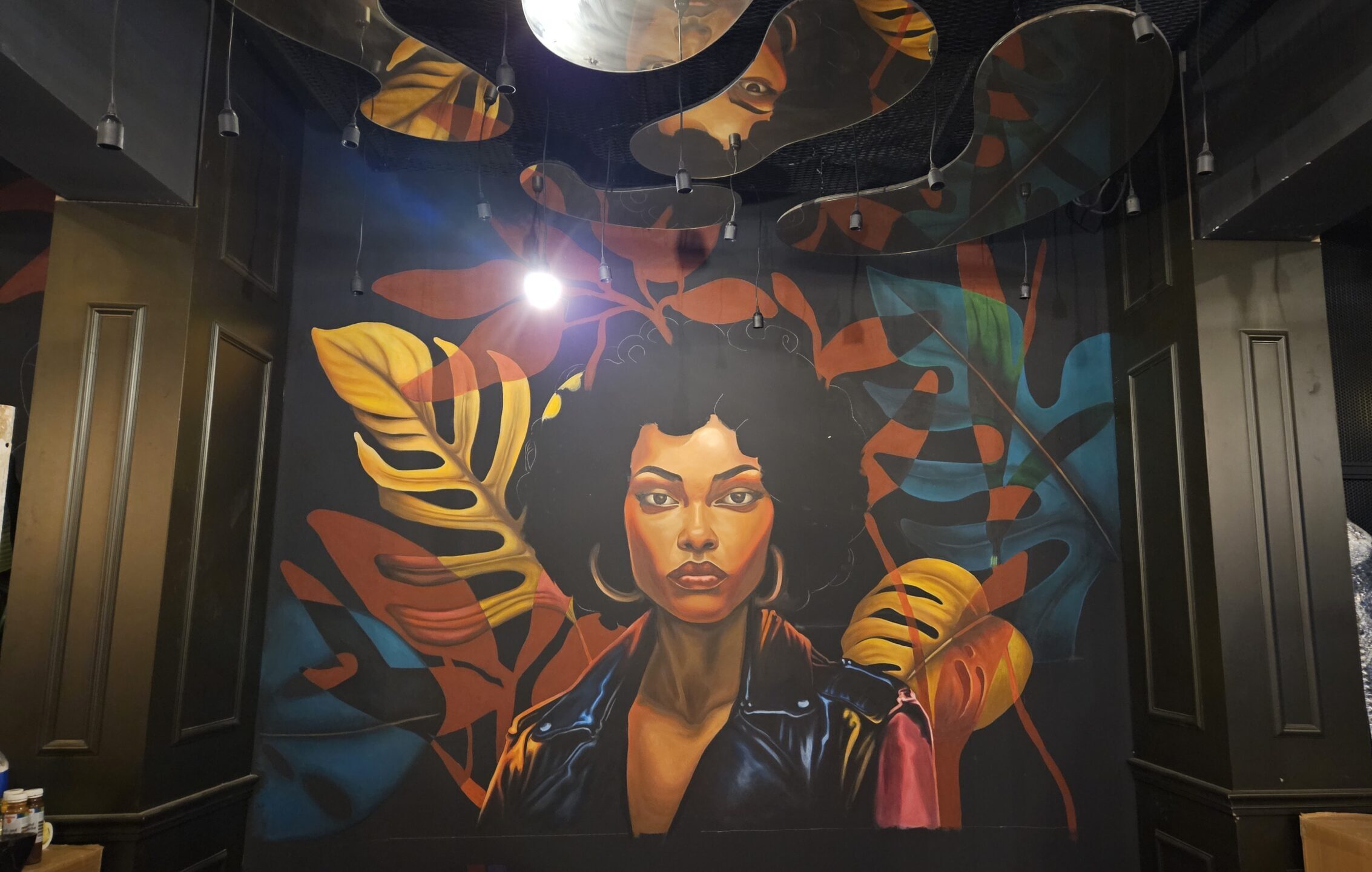 Indoor mural painting in Berlin café restaurant featuring a powerful portrait of a Black woman with afro hair and a leather jacket, surrounded by vibrant tropical leaves. Wall art by Berlin-based mural artist Cemre Şenoğlu Krahn.